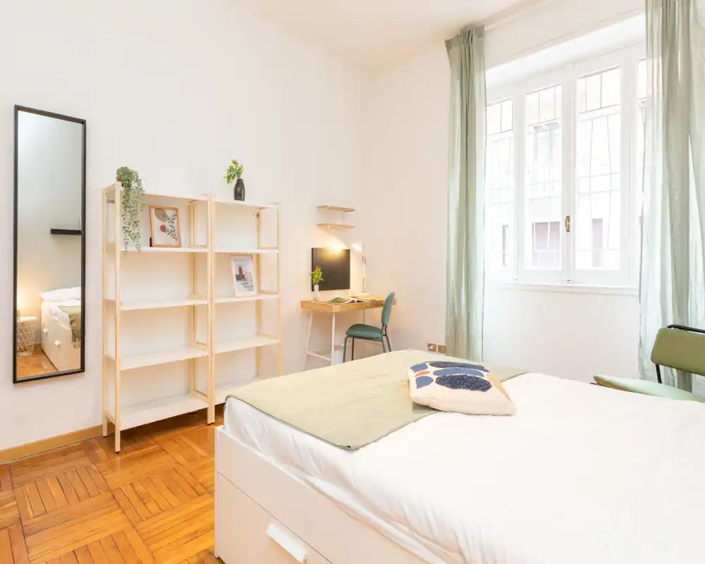 Bright, modern bedroom with a neatly made double bed in the foreground, a wooden parquet floor, open shelving, a small desk and large window letting in ample natural light.