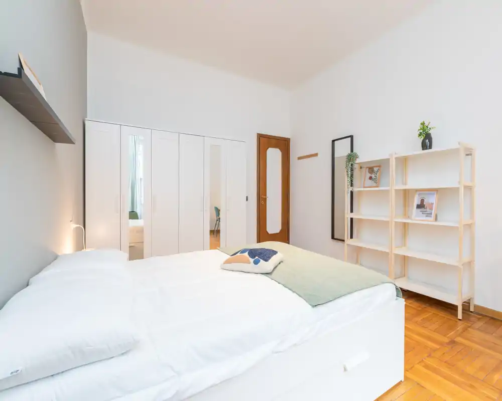Bright, minimal bedroom with a large white bed, wooden parquet floor, wardrobe with mirrored doors and open shelving. Clean styling and soft textiles create a calm, modern atmosphere ideal for rental listings.