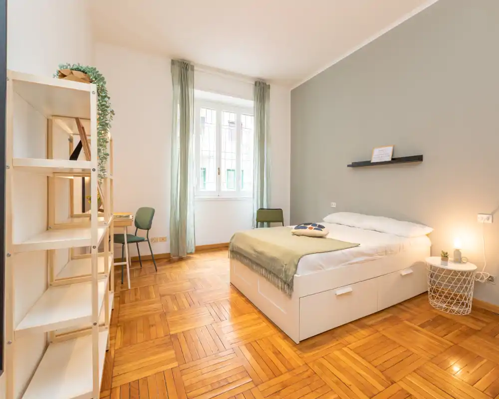 Bright, modern bedroom with a comfortable double bed, soft green accents and large window providing natural light. The room features parquet flooring, minimalist shelving and a small desk area — ideal for relaxing or working.