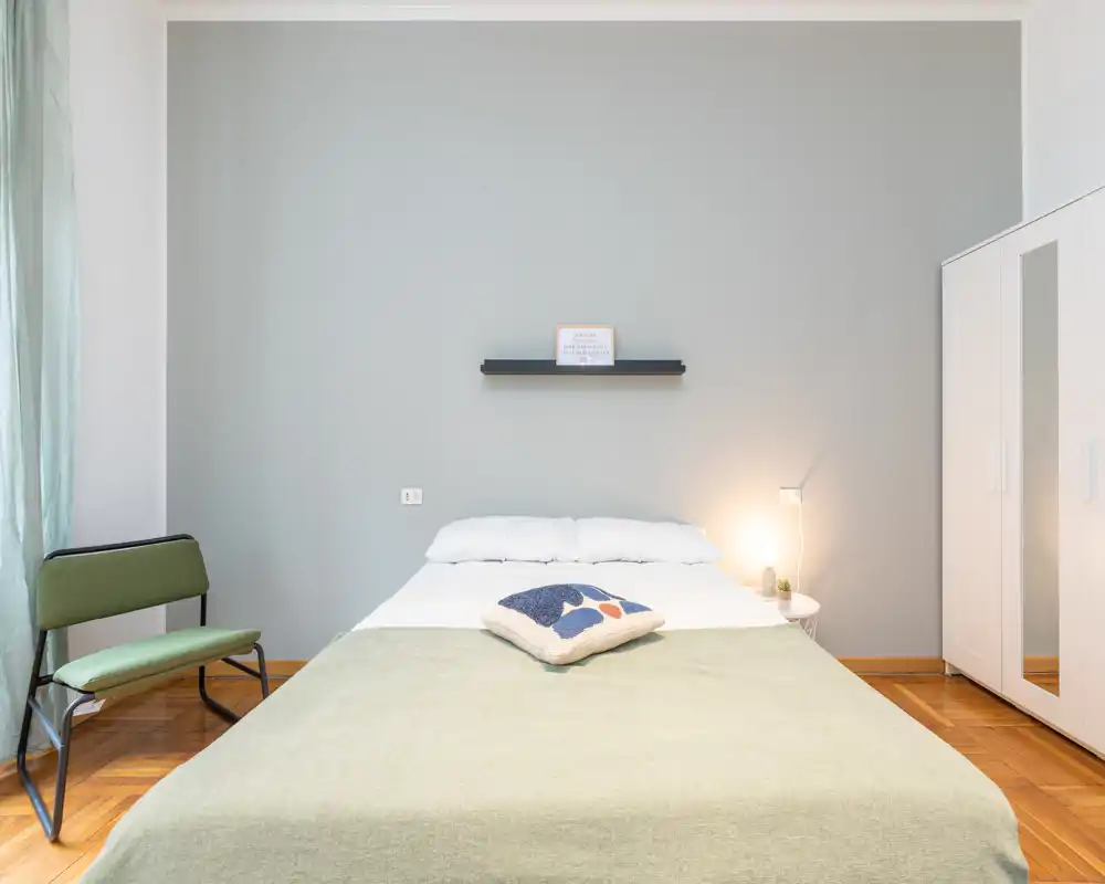 Bright, minimalist bedroom with a centrally placed double bed, soft linens, a decorative cushion and a bedside lamp; clean parquet floor and a mirrored wardrobe add function and light.