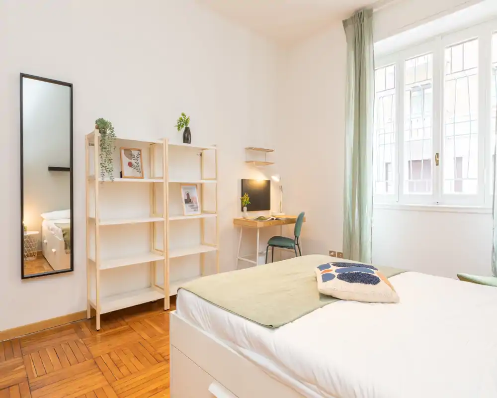 Bright, well-staged bedroom with a comfortable double bed, soft textiles and a small workspace by the window. Natural light fills the room and simple wooden shelving and mirror add a clean, modern touch.