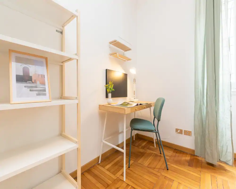 Bright and minimal living area with a compact wooden desk and chair by the window, tasteful shelving and warm parquet flooring — a calm workspace corner inside a small living room.