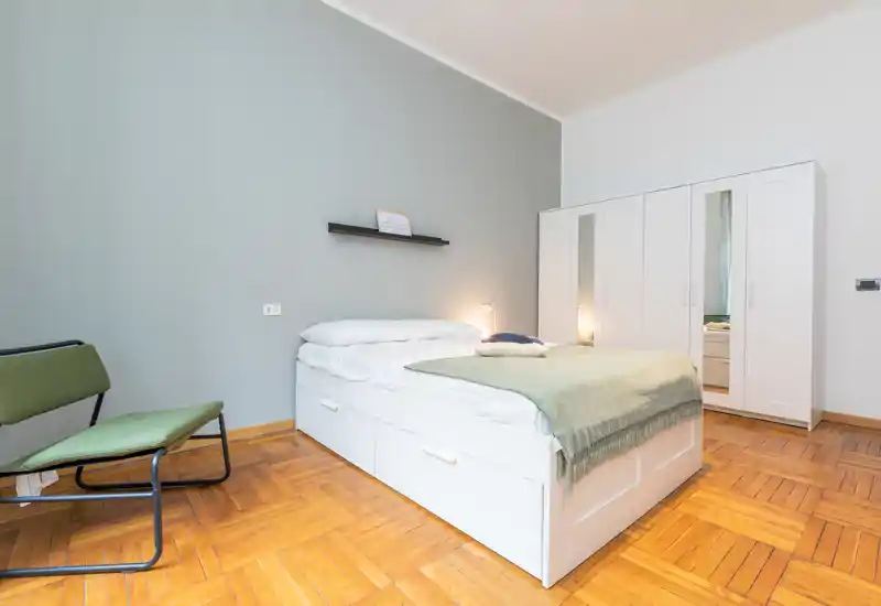 Bright, modern bedroom with a large white platform bed, cozy green throw and minimalist wardrobe; warm parquet flooring and simple decor create a calm, inviting space.