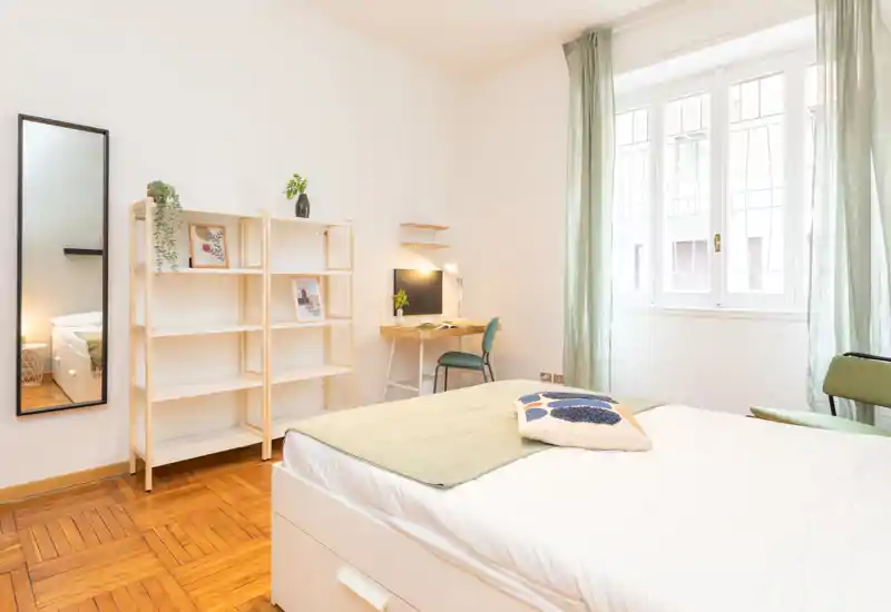 Bright, modern bedroom with a neatly made double bed in the foreground, a wooden parquet floor, open shelving, a small desk and large window letting in ample natural light.