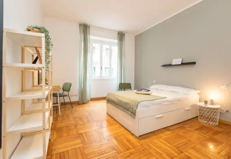 Bright, modern bedroom with a comfortable double bed, soft green accents and large window providing natural light. The room features parquet flooring, minimalist shelving and a small desk area — ideal for relaxing or working.