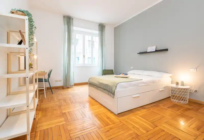 Bright, well-staged bedroom with a double bed, bedside lamp and soft textiles. Large window provides abundant natural light and warm parquet flooring enhances the cozy atmosphere.