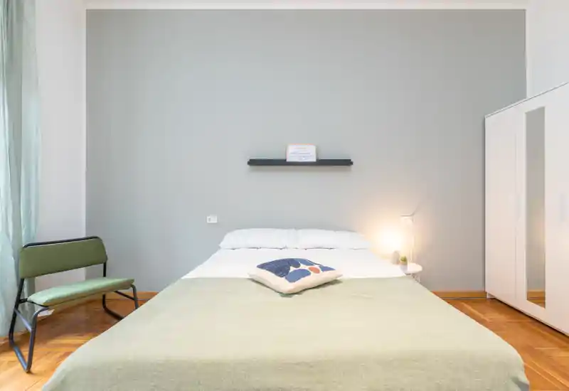 Bright, minimalist bedroom with a centrally placed double bed, soft linens, a decorative cushion and a bedside lamp; clean parquet floor and a mirrored wardrobe add function and light.