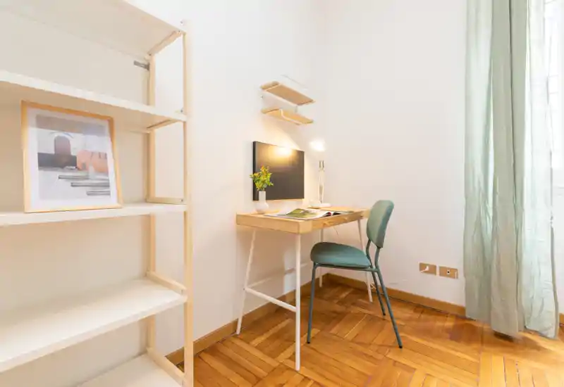 Zona giorno luminosa e minimale con una scrivania compatta in legno e sedia vicino alla finestra, scaffalatura estetica e pavimento in parquet caldo — un angolo lavoro tranquillo all'interno di un piccolo soggiorno.