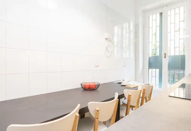 Angolo cucina luminoso e minimale con un lungo tavolo/bancone e quattro sedie, luce naturale dalle porte-finestre alte e una ciotola di frutta semplice sul tavolo.