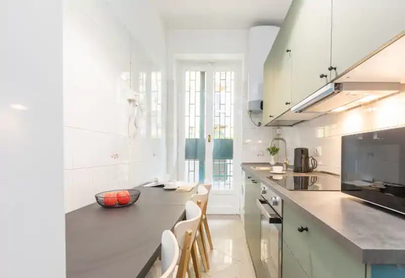 Cucina moderna a corridoio luminosa con mobili verdi, forno e piano cottura integrati e un bancone pranzo compatto per tre persone — ben illuminata da una finestra alta.