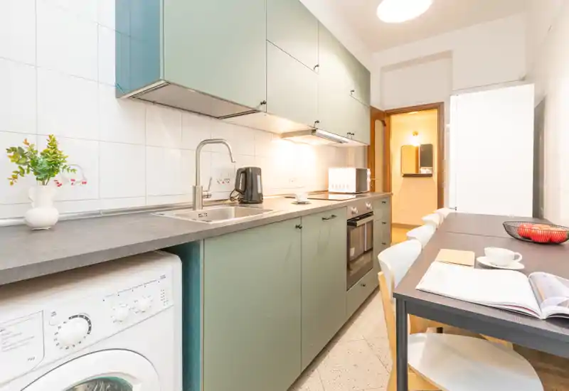 Cucina moderna e luminosa con mobili verdi, forno integrato e lavatrice, piccolo tavolo da pranzo apparecchiato e accessori sul piano di lavoro.