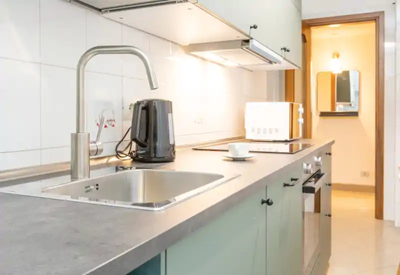 Cucina moderna e luminosa con lavello in acciaio, forno incassato, bollitore elettrico e microonde — piano di lavoro pulito e mobili verdi chiaro.
