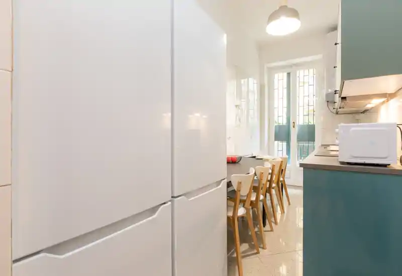 Cucina luminosa con frigorifero in primo piano, tavolo lungo con sedie e accesso a una porta/balcone che garantisce luce naturale.