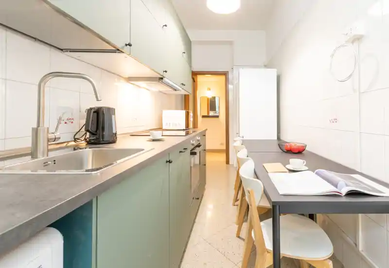 Cucina a corridoio moderna con lungo piano di lavoro, lavello, forno incassato e tavolo stretto con sedie — luminosa e ben allestita per l'uso quotidiano.