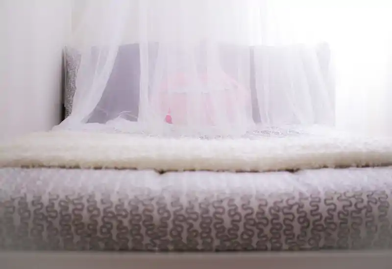 Bright, cozy bedroom close-up with a soft canopy over the bed, textured bedding and a decorative cushion. The image highlights a welcoming sleeping area with delicate textiles and natural light.