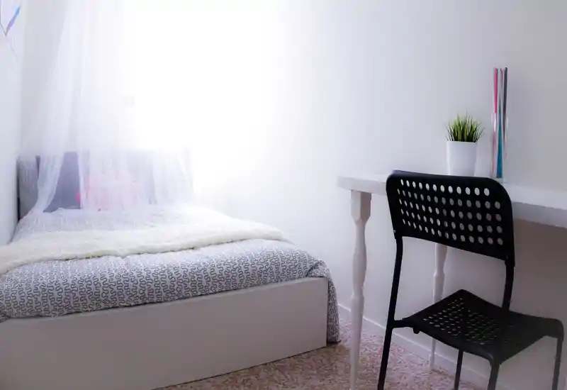 Bright, minimalist single bedroom with a neatly made bed, soft throw and delicate canopy; a compact desk and chair create a functional work corner.