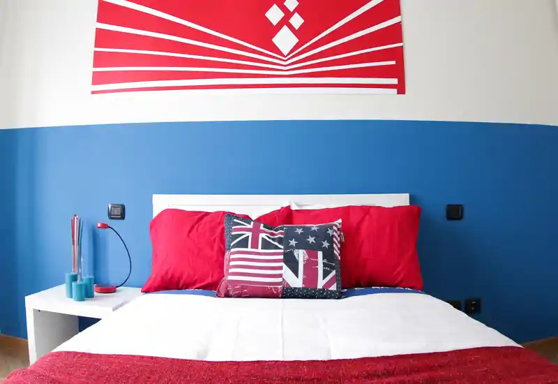 Bright, modern bedroom with a white headboard, red pillows and decorative Union-Jack themed cushions; clean white bedding and bold blue accent wall create a fresh, stylish look.