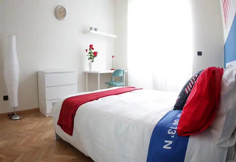 Bright, minimal bedroom with a neatly made double bed, red accents and a small desk by a large window. Clean parquet flooring and simple white furniture create a fresh, airy feel ideal for rental listings.