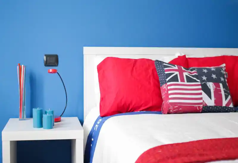 Bright, modern bedroom corner with a white headboard, vivid red pillows and a decorative flag-pattern cushion. A minimalist white bedside table with blue candles and a slim red lamp complements the bold blue wall.