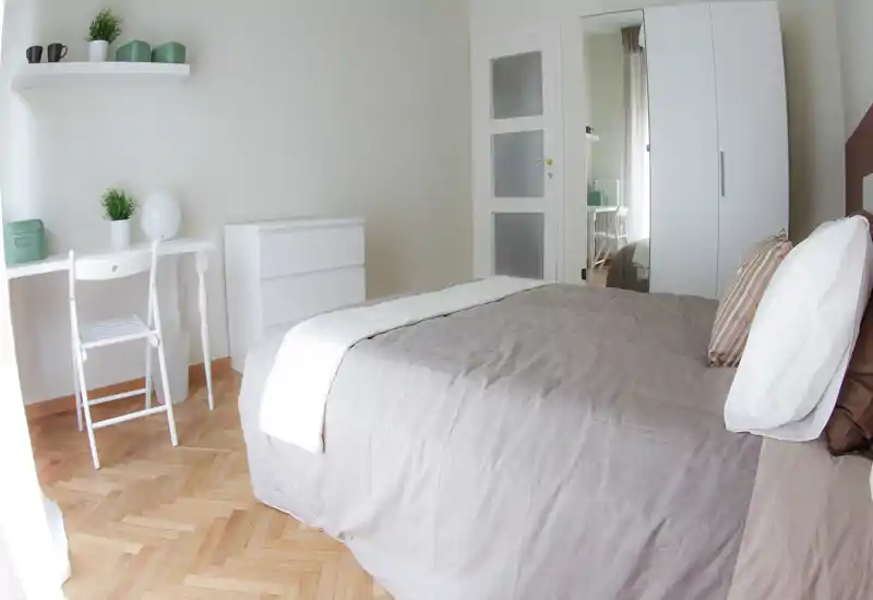 Bright, modern bedroom with a neatly made double bed, wooden parquet flooring and minimalist white furniture. Large wardrobe with mirrored door and a small desk make this room practical and airy.