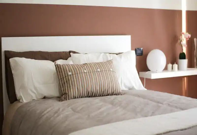 Tastefully styled bedroom with a comfortable double bed, layered linens and a decorative striped cushion. Warm earth-toned wall and bedside shelf create a cozy, well-lit atmosphere ideal for listing photos.