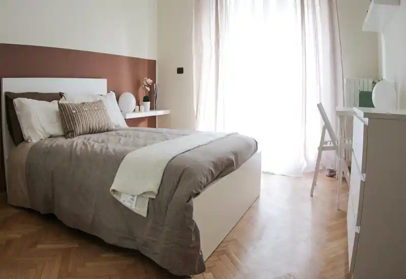 Bright, cozy bedroom with a neatly made double bed, soft linens and natural light from a large window.