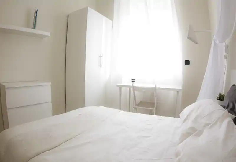 Bright, minimal bedroom with white linens, a wardrobe and a small desk by a large window — clean modern styling and soft natural light.