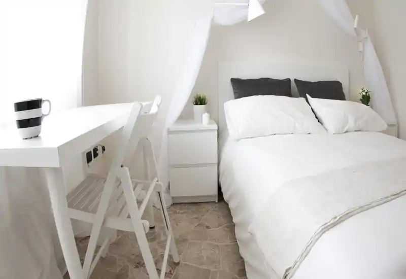Bright, minimalist bedroom with a neatly made double bed, light textiles and a small bedside table with decorative plants. The clean white palette and canopy create an airy, inviting atmosphere ideal for rentals.