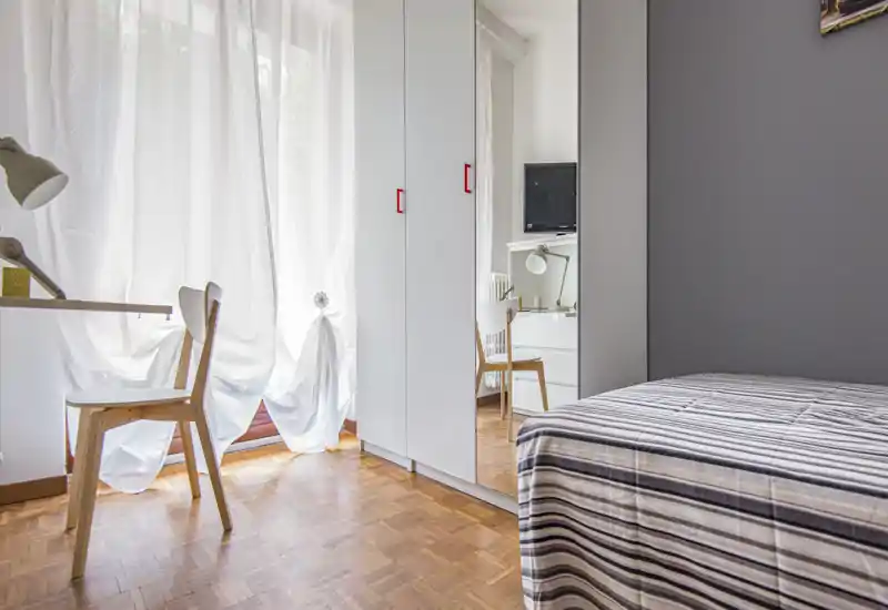 Camera da letto luminosa, arredata con un letto singolo a righe, armadio con anta specchiata e una piccola scrivania vicino alla finestra con tende leggere; ambiente pulito e moderno.