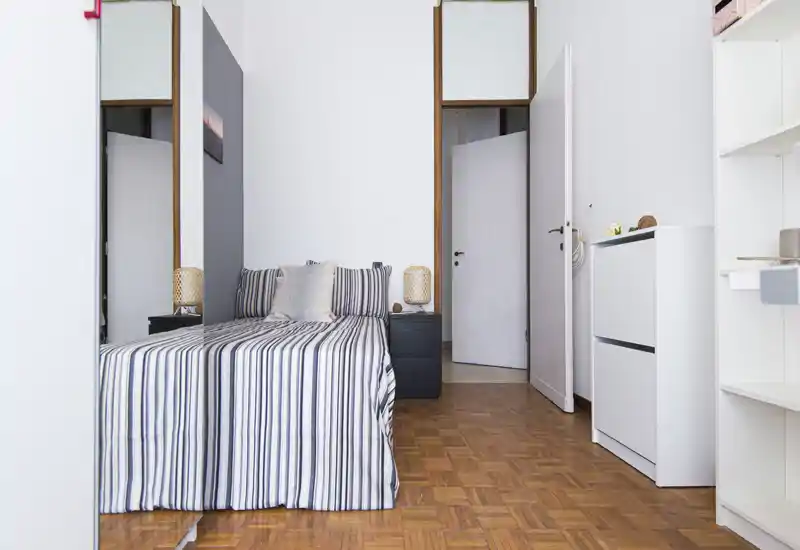 Camera da letto luminosa e ordinata con letto matrimoniale con biancheria a righe, comodini con lampade e pavimento in parquet chiaro — perfetta per annunci in affitto che evidenziano uno spazio notte accogliente e pulito.