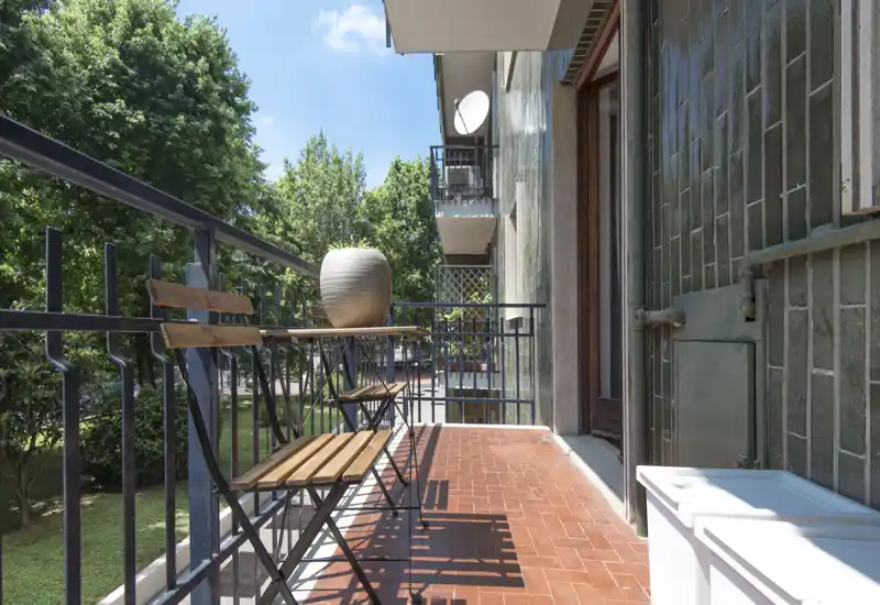 Balcone soleggiato con piccolo tavolo e sedie in legno, pavimento in cotto e vista su alberi — un angolo accogliente per il caffè del mattino.