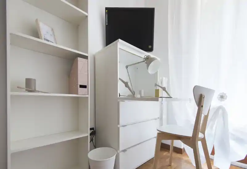 Angolo camera luminoso e minimale con cassettiera bianca, lampada da scrivania e una sedia semplice accanto a tende leggere — un piccolo spazio di lavoro dallo stile nordico.