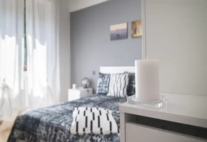 Bright, modern bedroom with patterned bedding, decorative cushions and natural light from a sheer-curtained window. The room features a bedside table and tasteful minimal decor for a calm atmosphere.