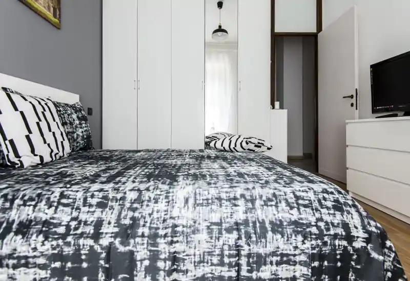 Camera da letto moderna e luminosa con un grande letto in primo piano, biancheria con motivo, armadio a muro bianco e comò con TV — pulita, ben illuminata e accogliente.