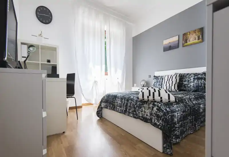 Camera da letto moderna e luminosa con letto matrimoniale, biancheria a motivo e grande finestra che illumina la stanza; scrivania e scaffali semplici per un ambiente funzionale e di design.