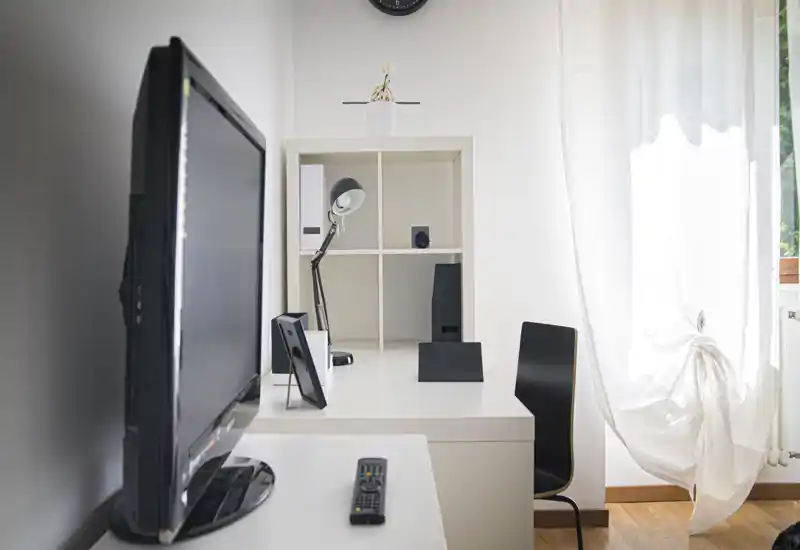 Bright, minimal living area with a TV, desk, shelving unit and a chair beside a sunlit window — clean, simple and functional.