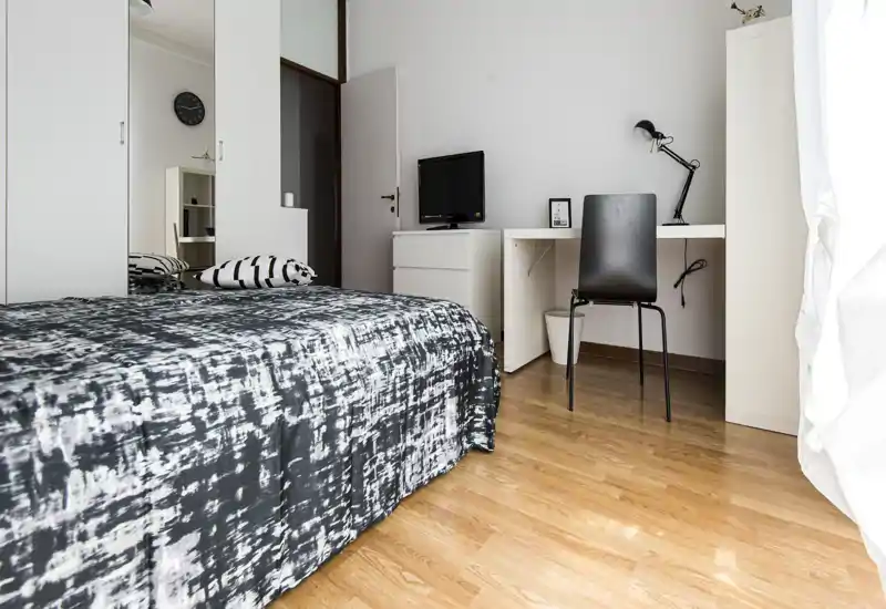 Camera da letto luminosa e minimale con letto matrimoniale con copriletto a fantasia, piccolo scrittoio con lampada e sedia, armadio con ante a specchio e pavimento in legno.