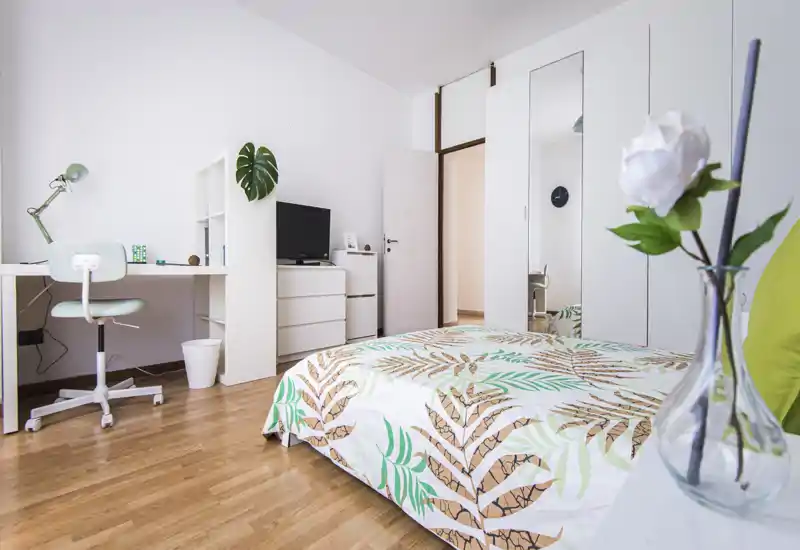 Camera da letto moderna e luminosa con copriletto a motivo, armadi bianchi e una piccola scrivania — ideale per un soggiorno confortevole e smart working.