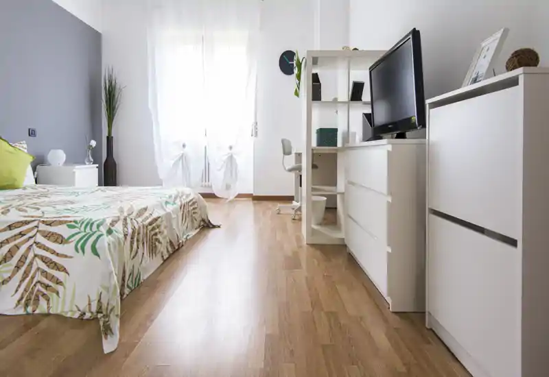 Camera da letto luminosa e moderna con copriletto a foglie, comodino e TV su cassettiera; luce naturale che entra dalle tende leggere.