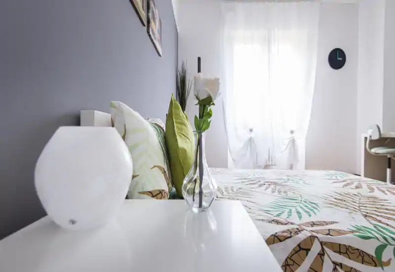 Camera da letto luminosa e ben arredata con biancheria a fantasia, cuscini decorativi e luce naturale da una tenda leggera. Il letto è il punto focale e l'ambiente risulta fresco e accogliente.