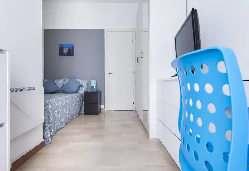 Angolo camera luminoso e moderno con letto dalla fantasia elegante, cuscini e un comodino con lampada. Pareti bianche pulite e un'area studio con sedia blu completano l'ambiente ordinato.