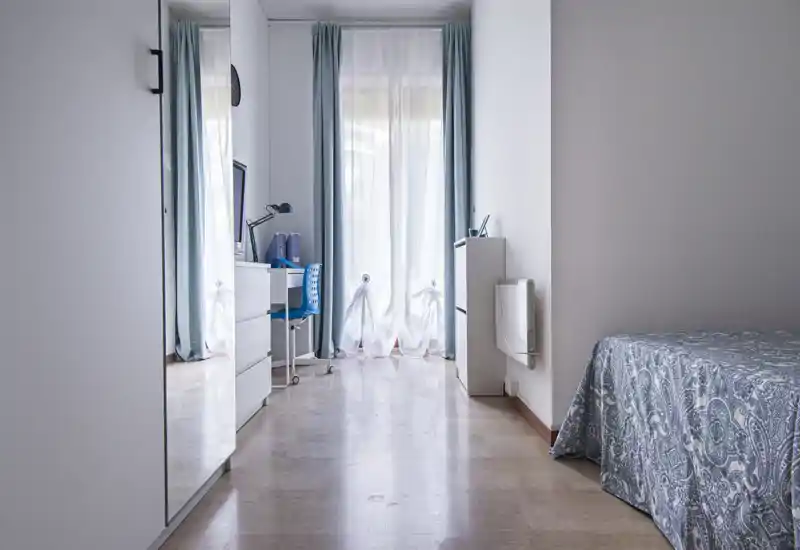 Angolo camera da letto luminoso e minimalista con letto parzialmente visibile coperto da una coperta stampata, scrivania e sedia accanto a una finestra con tende leggere che lasciano entrare luce naturale.