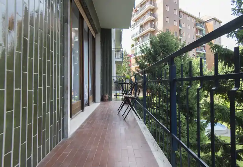 Balcone cittadino soleggiato con pavimento in piastrelle, ringhiera metallica e un piccolo tavolo da bistrò; affaccia su alberi e palazzi vicini, perfetto per il caffè mattutino.
