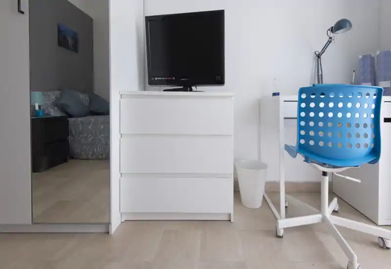 Angolo di camera da letto luminoso e moderno con area letto visibile a sinistra, comò bianco con TV e una piccola scrivania bianca con sedia girevole blu. L'immagine mostra un layout pulito e minimale, ideale per un annuncio di affitto.