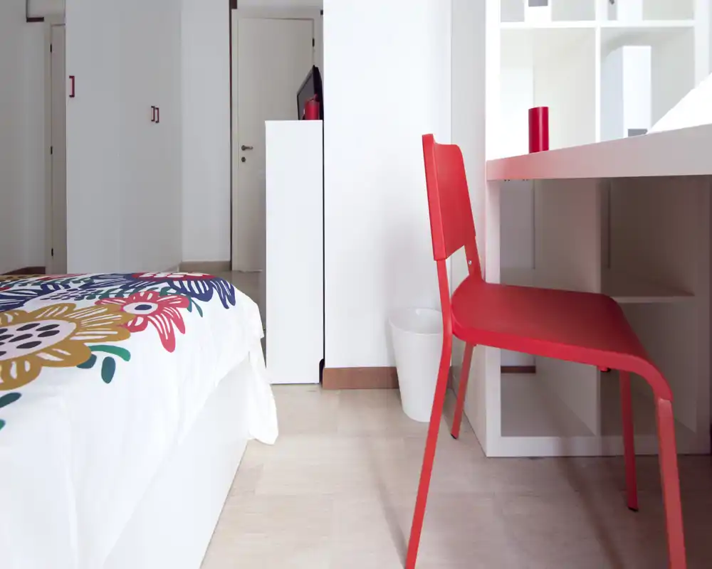 Angolo camera luminoso e moderno con una coltre floreale colorata e una sedia da scrivania rossa. La foto mette in evidenza un'area letto ordinata e una postazione compatta, ideale per una persona.