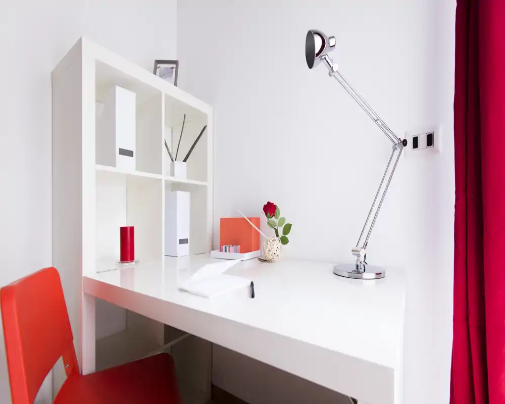 Angolo lavoro luminoso e moderno con scrivania bianca, lampada regolabile cromata, scaffalatura e dettagli rossi; perfetto per uno smart working compatto.