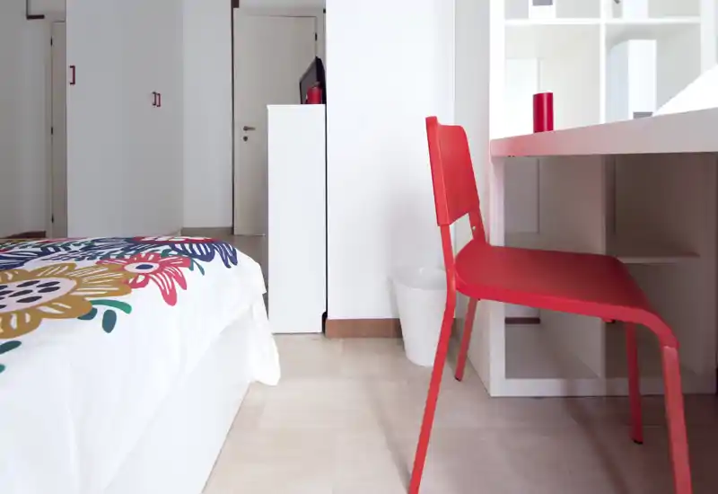Angolo camera luminoso e moderno con una coltre floreale colorata e una sedia da scrivania rossa. La foto mette in evidenza un'area letto ordinata e una postazione compatta, ideale per una persona.