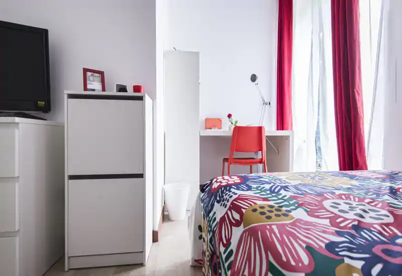 Camera da letto luminosa e colorata con copriletto a fantasia, scrivania e sedia vicino a una grande finestra che lascia entrare luce naturale — uno spazio notte ben curato e compatto.