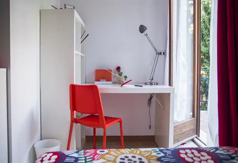 Angolo camera da letto con scrivania bianca, sedia rossa e copriletto colorato; luce naturale dalla porta del balcone che crea un'atmosfera fresca e accogliente.