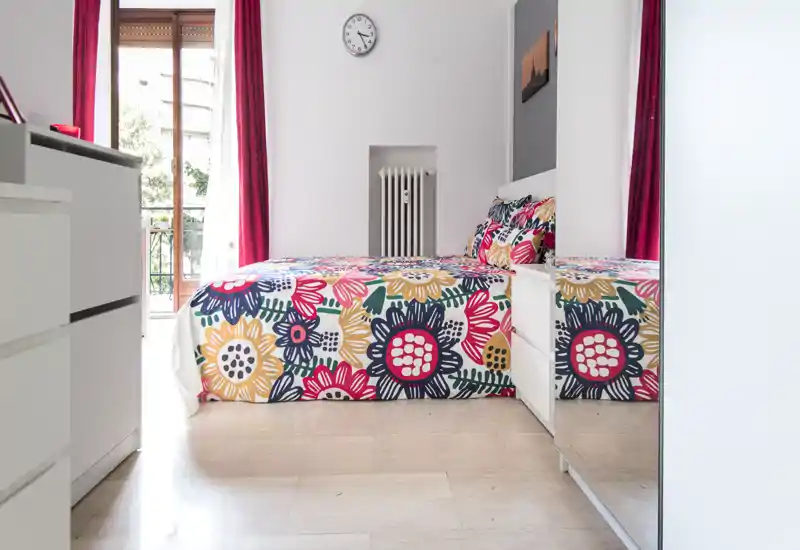 Camera da letto luminosa e ben curata con una coperta floreale colorata, luce naturale dal balcone e arredi moderni e puliti — perfetta per annunci che vogliono mostrare comfort e stile.